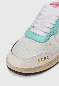 Bijela tenisica s tirkiznim akcentom, perforiranim prednjim dijelom i zlatnim "ACBC" logotipom. Teksturirana potplata s crnim detaljem na rubu.