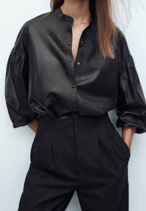 Frau trägt ein schwarzes Lederhemd mit Knöpfen und 3/4-ärmeln mit Puffeffekt, das in hoch taillierte, schwarze, plissierte Hosen gesteckt ist, mit Händen in den Taschen.
