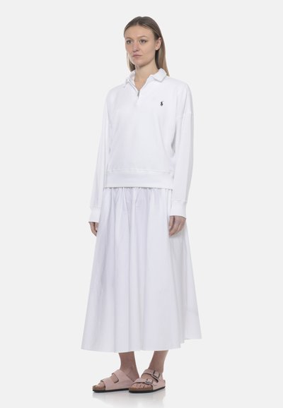 Donna che indossa una maglia bianca a maniche lunghe con colletto con cerniera, una gonna bianca alla caviglia e sandali beige con due fibbie, in piedi su uno sfondo semplice.