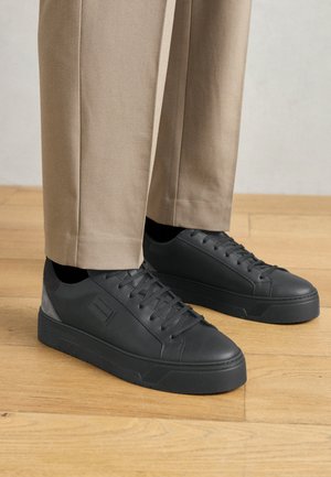 Sneakers neri con tomaia in pelle liscia, dettagli grigi texturizzati e suola in gomma spessa. Indossati con pantaloni beige chiaro.