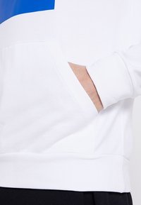 Sweat-shirt blanc avec un accent bleu, doté d'une poche avant et d'un ourlet côtelé. Tissu doux avec une texture lisse.