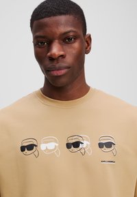 Beige környakú póló, amelyen négy grafikai illusztráció látható egy napszemüveget viselő karakterről fekete-fehérben, az "KARL LAGERFELD" felirattal.