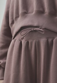 Sudadera púrpura con textura acanalada, que presenta una cintura con cordón y tela fruncida debajo. Ajuste suave y ligeramente holgado.