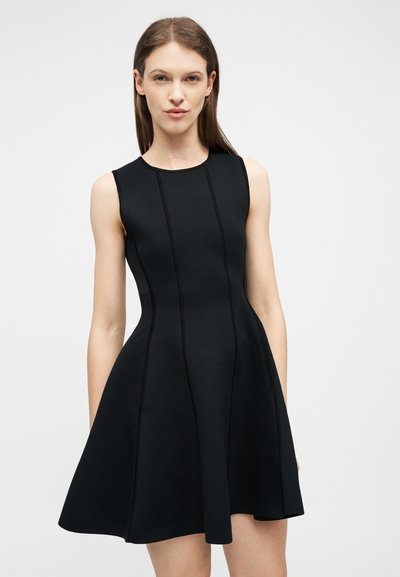 Γυναίκα που φοράει μαύρο αμάνικο φόρεμα σε σχήμα Α με κατακόρυφες ραφές, στέκεται μπροστά σε απλό ανοιχτό γκρι φόντο.