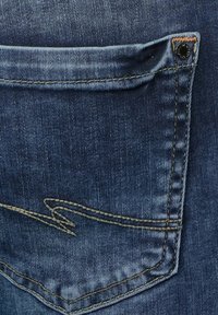 Tmavě modré džínové kalhoty s zadní kapsou, která má mírně složenou klopu, černou nýtkou a kontrastním šitím v světle hnědé barvě.