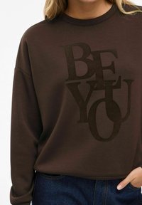 Sweatshirt marron avec un imprimé texturé noir portant l'inscription "SOIS TOI." Présente une coupe décontractée et un col rond, associé à un jean en denim.