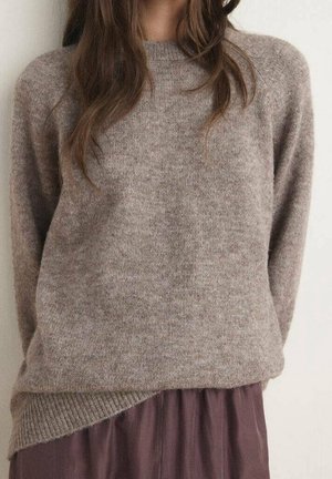 Kobieta ubrana w miękki, taupe sweter z dzianiny z prążkowanym wykończeniem u dołu, na luźnej, fiołkowej spódnicy, stojąca na jasnym tle.