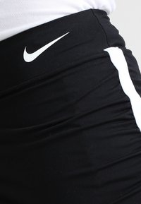 Czarne szorty sportowe z białymi paskami po bokach i małym białym logo Nike na pasku. Materiał wydaje się elastyczny i gładki.