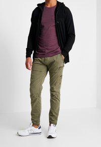 Sudadera negra con cremallera, camiseta de cuello redondo morada y pantalones cargo oliva con bolsillos. Zapatillas blancas con detalles azules completan el conjunto.