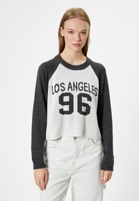 Grauer und weißer, kurz geschnittener Pullover mit Raglanärmeln, der "LOS ANGELES 96" in fettem schwarzem Text zeigt, mit Stricktextur und einfachem Design.