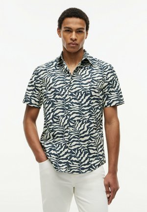 Uomo con camicia a maniche corte con bottoni a fantasia bianca e nera e pantaloni bianchi, in piedi davanti a uno sfondo bianco semplice.