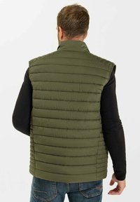 Grön pufferväst med hög krage, quiltad textur och ärmlös design. Bärs över en svart långärmad tröja och blå jeans.