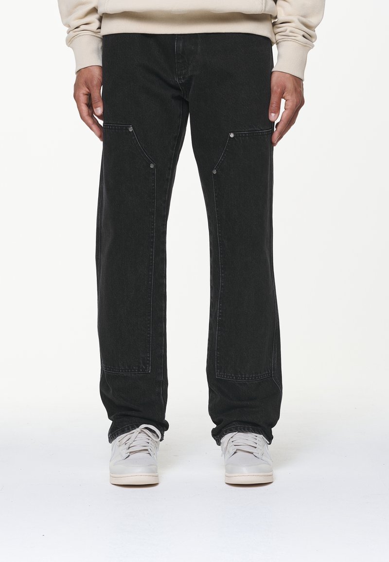 Personne portant un jean noir avec des poches plaquées à l'avant et des baskets blanches, debout devant un fond blanc uni.