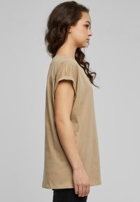 Beige bomulls T-shirt med rullade ärmar, avslappnad passform, rund halsringning. Mjuk textur, rak nederkant, bärs med svarta byxor. Sidovy.