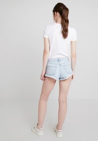 Mujer con cabello castaño en una coleta, vistiendo una camiseta blanca, pantalones cortos de mezclilla azul claro y zapatillas blancas Reebok, de pie mirando hacia atrás.