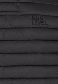 Schwarzer, gesteppter Stoff mit horizontalen Nähten und schwarzem, gesticktem Text "SUB LEVEL" im oberen rechten Abschnitt.