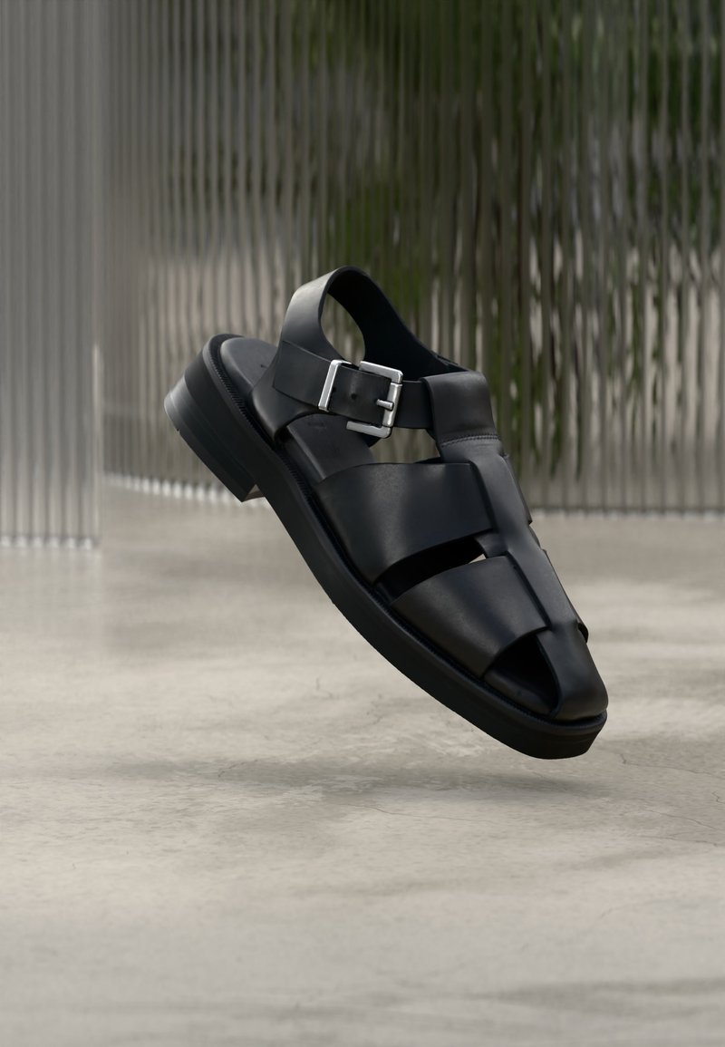 Μαύρο δερμάτινο ανδρικό ψαράδικο σανδάλι με λουράκι με πόρπη, ανοιχτά δάχτυλα και χαμηλό τακούνι, εμφανίζεται πάνω σε ουδέτερο σύγχρονο φόντο.