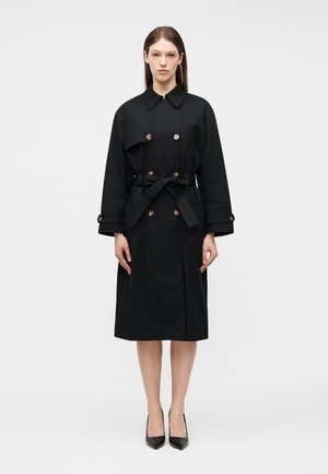 Γυναίκα που στέκεται φορώντας ένα μαύρο καμπαρντίνα μήκους μέχρι τα γόνατα με ζώνη δεμένη στη μέση και μαύρα ψηλοτάκουνα παπούτσια σε απλό φόντο.