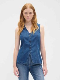 Woman with long red hair wearing a sleeveless blue denim buttoned vest and blue jeans, standing against a plain light background.