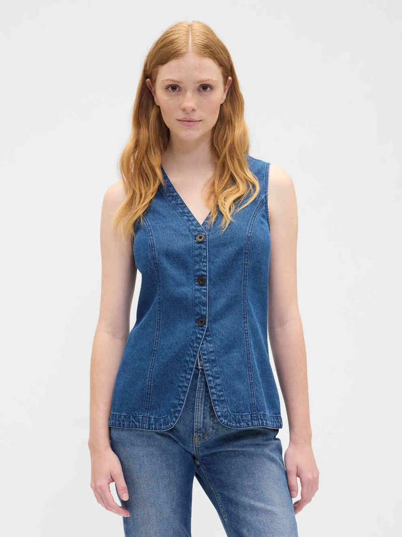 Woman with long red hair wearing a sleeveless blue denim buttoned vest and blue jeans, standing against a plain light background.