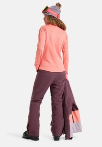Light pink long-sleeve top, purple snow pants, gray and orange jacket, knit lavender hat, holding jacket, black boots, textured materials.