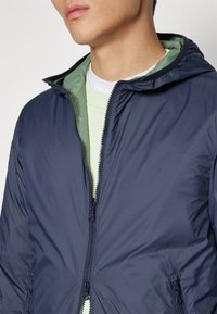 Marineblå hettegenser med glidelås og elastiske mansjetter. Fôret med lysegrønn og laget av glatt, vannavstøtende stoff.