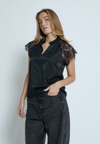 Sort bluse med blonder korte ærmer, nøglehulhalsudskæring og flæsekrave. Båret med mørkegrå højtaljede denimbukser.