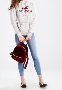 Šedá mikina s červeným logem "Hollister", modré slim džíny, černé balerínky a burgundský sametový batoh s přední kapsou a zipem.