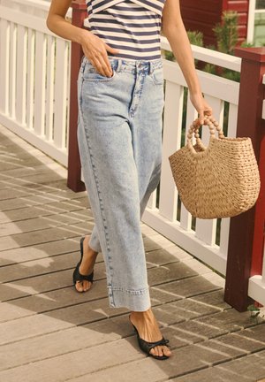 Osoba koja nosi prugastu majicu i svijetloplave traperice, drži ispleteni ručni torbu, hoda drvenom terasom blizu bijele ograde.