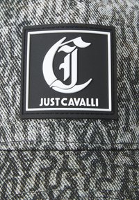 Parche con el logo de Just Cavalli en blanco y negro sobre un fondo de tela texturizada en gris y negro.