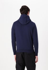 Homme portant un sweat à capuche bleu marine et un pantalon noir, debout face à un mur blanc, dos à la caméra.