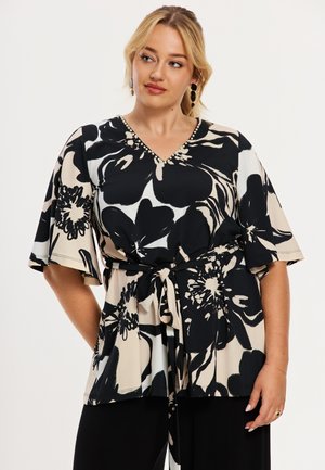 Kvinde med blondt hår iført sort og beige blomstret V-hals top med vide ærmer og bundet taljebælte, kombineret med sorte bukser, ser til siden.