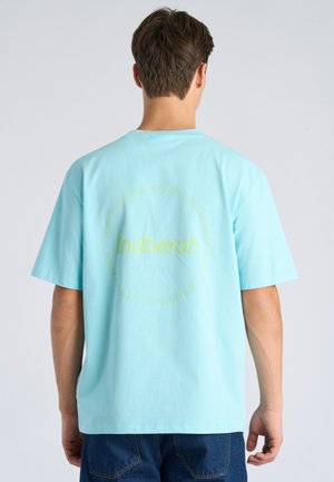 Man draagt lichtblauw T-shirt met gele cirkelvormige tekst en logo op de achterkant, gecombineerd met donkerblauwe spijkerbroek, staand tegen een effen achtergrond.