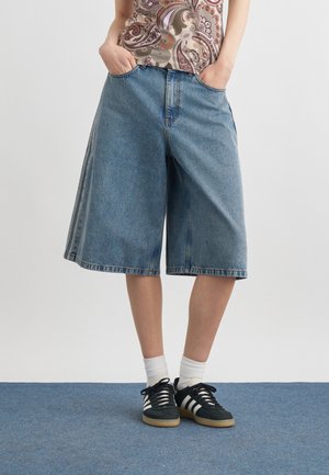 Person iført løstsiddende, knælange denimshorts, en mønstret top, hvide sokker og sorte Adidas-sneakers med hvide striber.