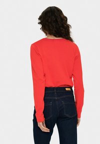 Raudona megztinis ilgomis rankovėmis su apvaliu iškirpimu, pagamintas iš minkštos medžiagos, derinamas su tamsiai mėlynomis džinsais, turinčiais kontrastingą oranžinę siuvimo nėrimą ir užpakalines kišenes.