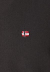 Pequeña bandera noruega bordada en rojo, blanco y azul sobre un fondo de tela de punto gris oscuro.