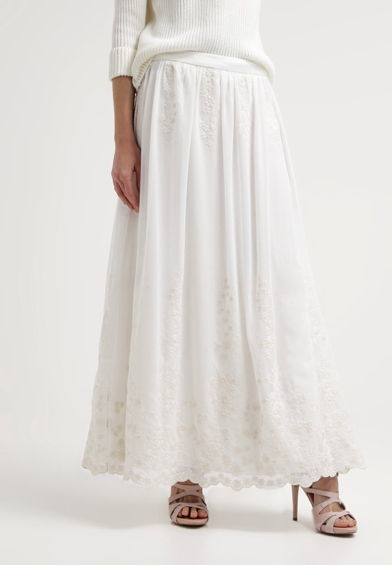 Jupe maxi blanche brodée portée avec un pull blanc et des sandales à talons beiges sur un fond uni.