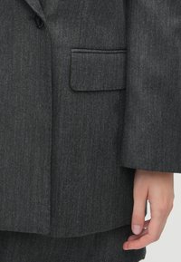 Blazer gri închis dintr-un material texturat, cu un singur nasture, buzunar cu clapetă și mâneci ușor supradimensionate, având detalii elegante de cusătură.