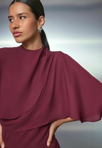 Rochie burgundă cu mâneci fluide și drapate și un guler înalt. Modelul poartă o cercei decorativ cu un design în formă de stea. Textură de țesătură netedă.