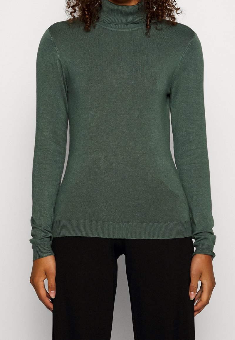 Personne portant un pull à col roulé vert à manches longues et un pantalon noir, debout devant un fond clair uni.