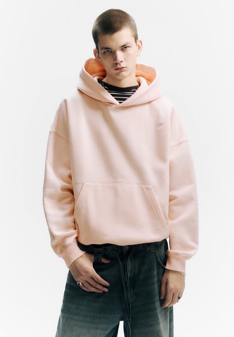 Sweat à capuche rose clair avec une poche frontale, associé à un jean gris ample. Comprend une doublure intérieure rayée dans la capuche.