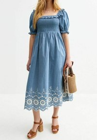 Denim midi šaty s nabíranými rukávy, pružným živůtkem, vyšívaným bílým květinovým vzorem na lemu, spárované s hnědými podpatkovými sandály a tkanou kabelkou.