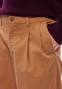 Barna, puha pamut keverék anyagból készült, pliszírozott nadrág, fém gombos záródással és oldalzsebekkel, sötétlila pulóverrel párosítva.