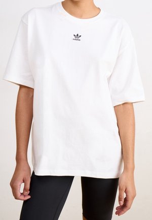Persona vistiendo una camiseta blanca oversize de Adidas con el logo negro en el pecho y pantalones cortos ajustados negros, de pie con los brazos relajados a los costados.