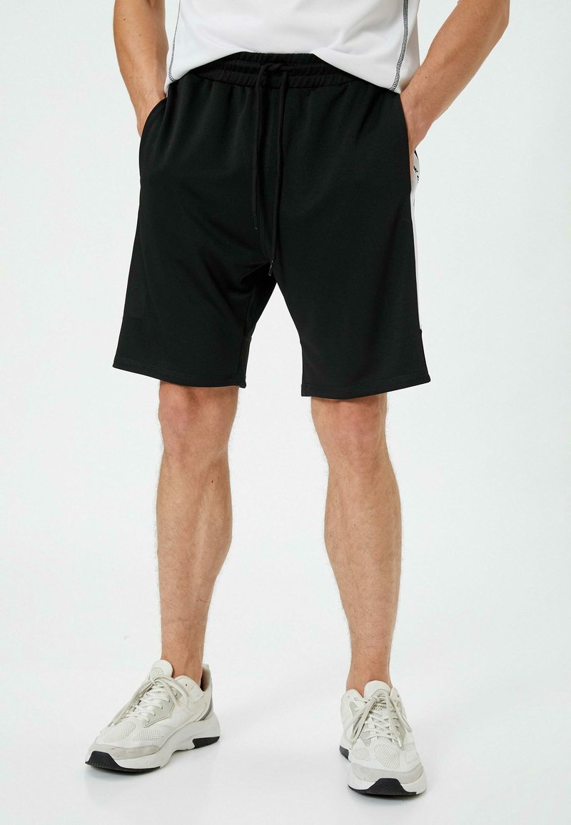 Sorte atletiske shorts med elastisk talje og snor, med sidelommer og en blød, glat overflade. Parret med hvide sneakers.