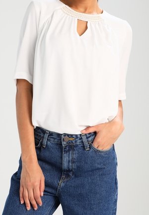 Blouse blanche à manches courtes avec détail de perles au col, associée à un jean en denim bleu taille haute, une main dans la poche.