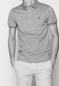 Randig polo-tröja i grått och vitt med en solid krage och tre-knappsstängning. Litet emblem på vänster bröst. Korta ärmar.