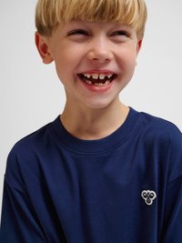 Ung dreng med blondt hår iført en mørkeblå skjorte, der smiler med synlige mellemrum i fortænderne mod en ensartet lys baggrund.