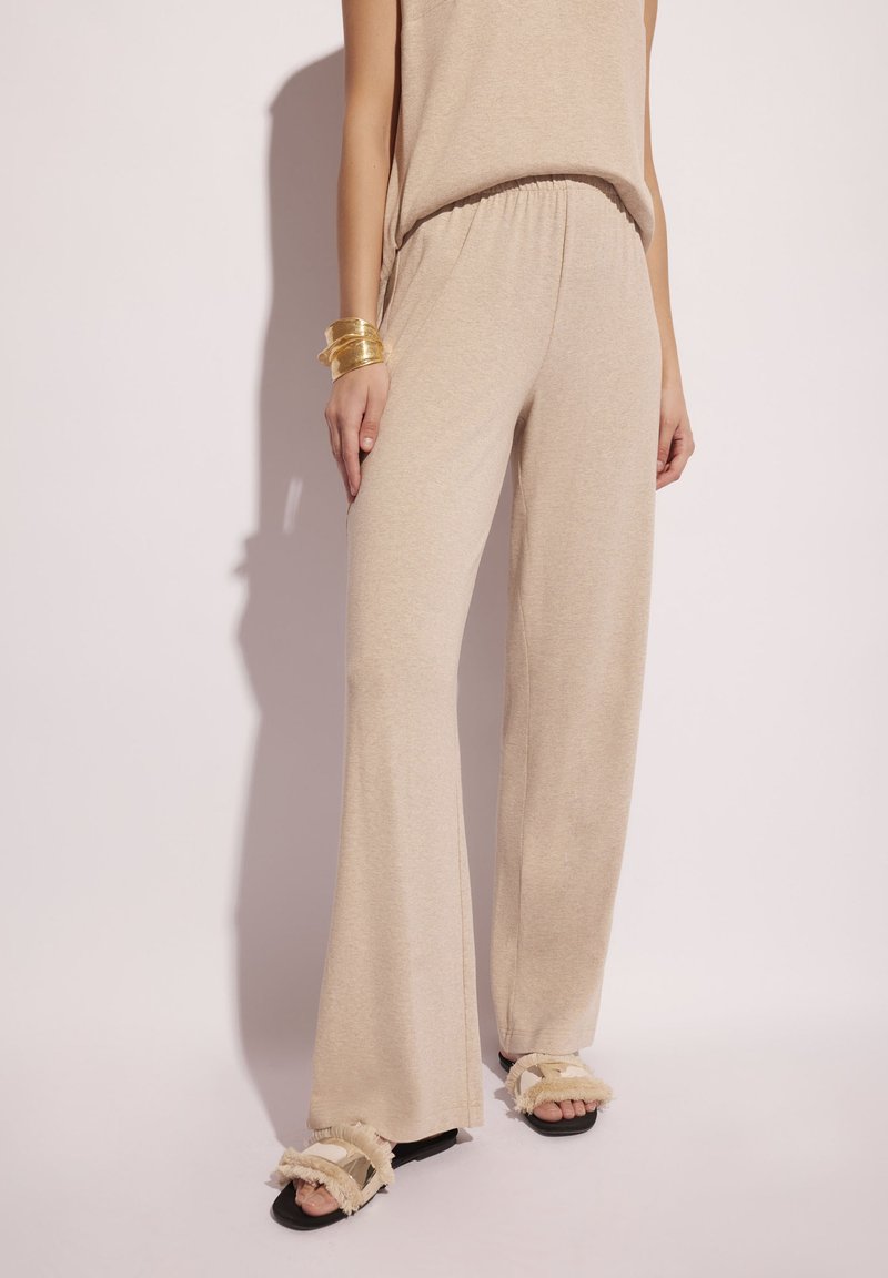 Beżowe spodnie dzwony z miękkiego materiału z elastycznym pasem, w połączeniu z czarnymi sandałami z frędzlami. Prosty design i faktura.