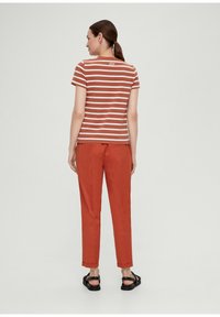 Gestreiftes T-Shirt in Braun und Creme mit rundem Ausschnitt, kombiniert mit orangen Hosen und schwarzen Sandalen. Das Shirt hat ein kleines Logo auf der Rückseite.